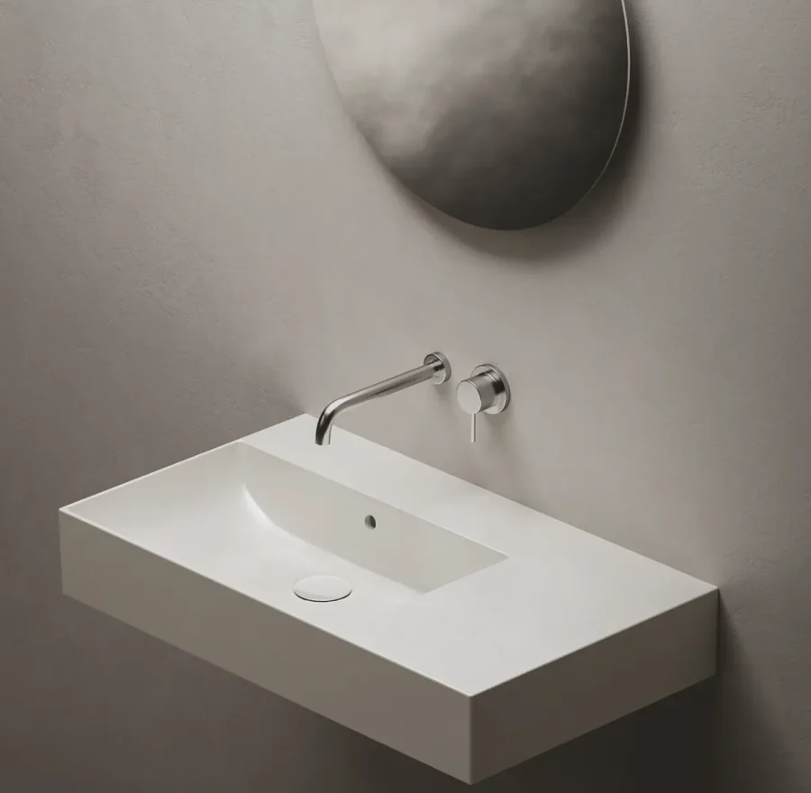 Lavabo sospeso bianco rettangolare con rubinetto a parete cromato, su parete grigio chiaro.