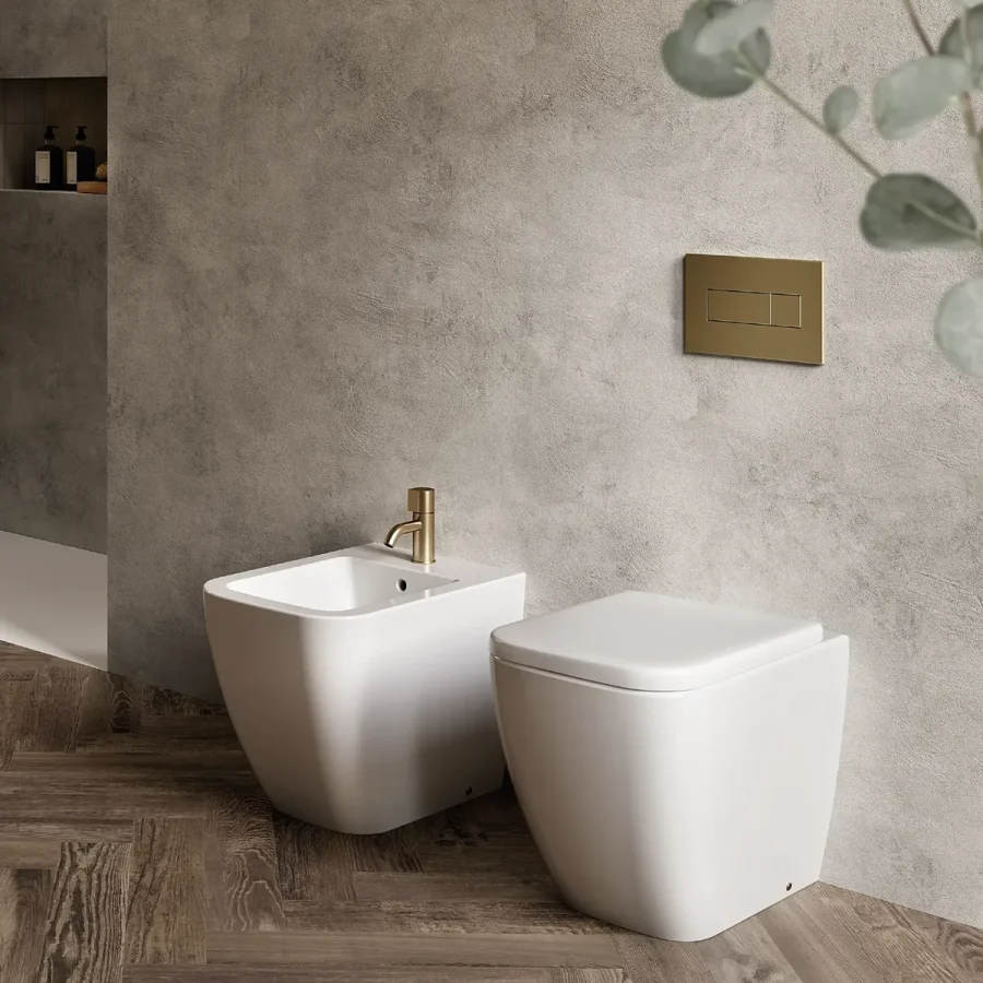 Coppia di sanitari bianchi a pavimento con rubinetto oro, su pavimento in legno e parete effetto cemento.