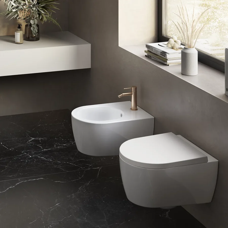 Coppia di sanitari sospesi bianchi con rubinetteria oro su pavimento in marmo nero e parete beige.