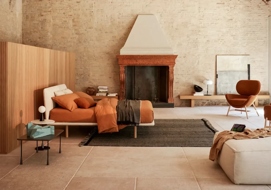 Letto con struttura minimal e testiera imbottita bianca in un ambiente con pareti in pietra e camino centrale.