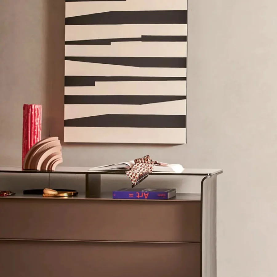 Cassettiera moderna con piano vetrato e oggetti decorativi, appoggiata a una parete chiara con quadro grafico