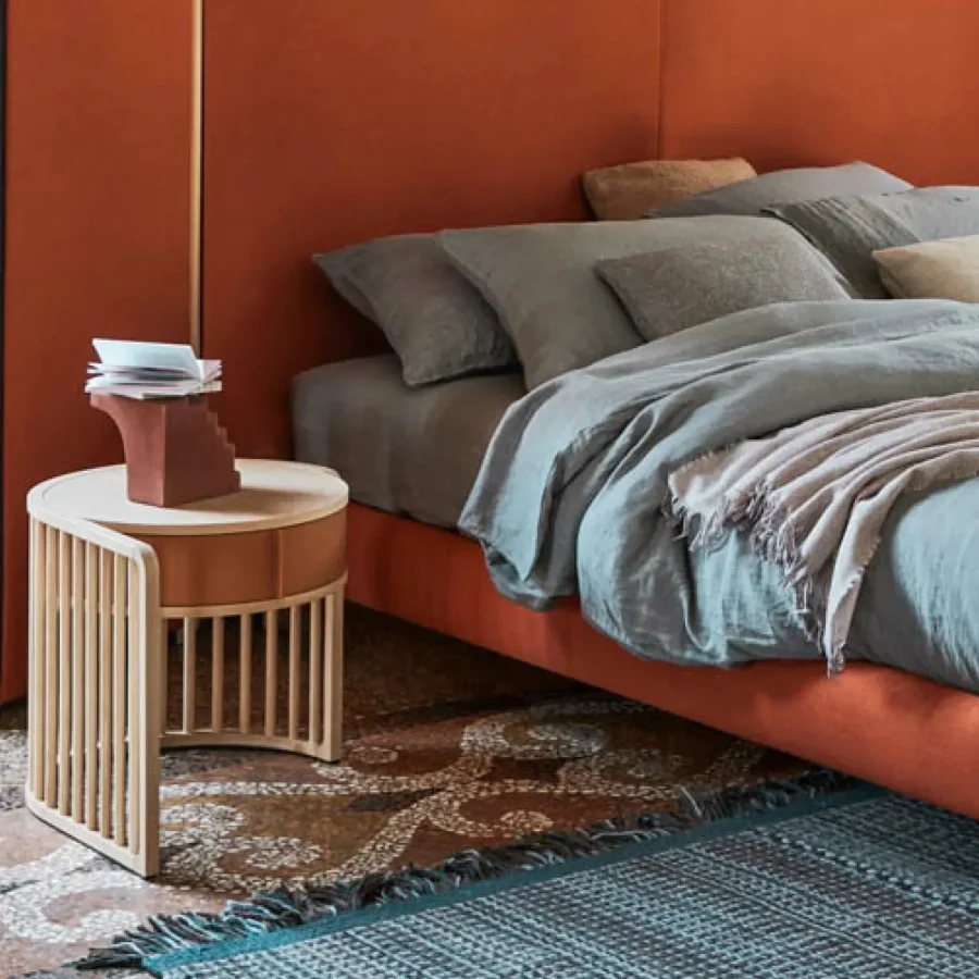 Comodino rotondo con struttura in legno chiaro e testiera del letto arancione sullo sfondo.