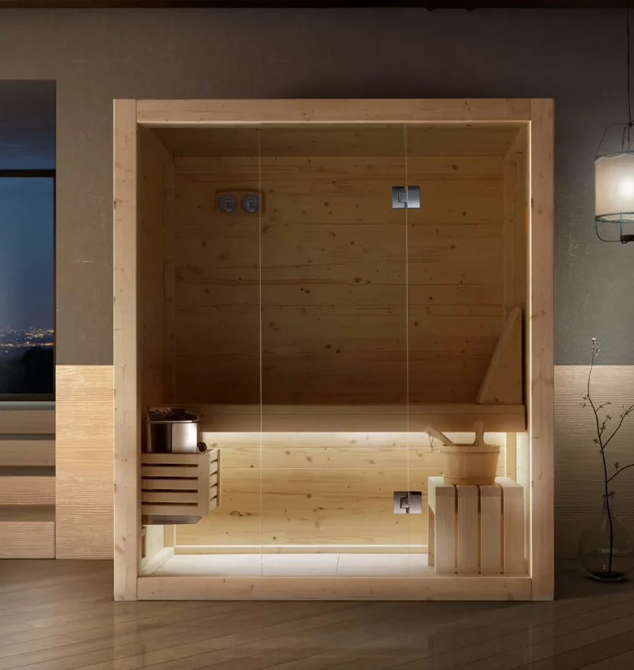 Sauna finlandese in legno chiaro con seduta interna illuminata e struttura in vetro, dotata di secchiello e mestolo in stile tradizionale.