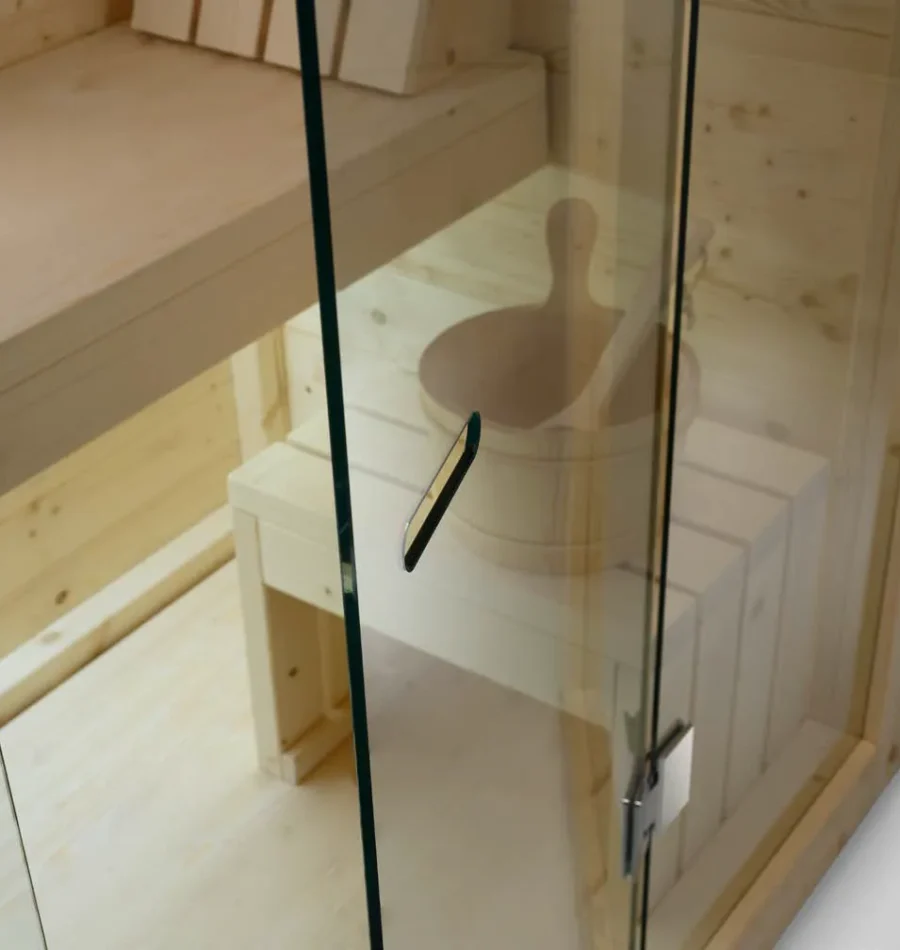 Dettaglio interno di sauna in legno con panca e secchiello tradizionale inquadrati da una porta in vetro.