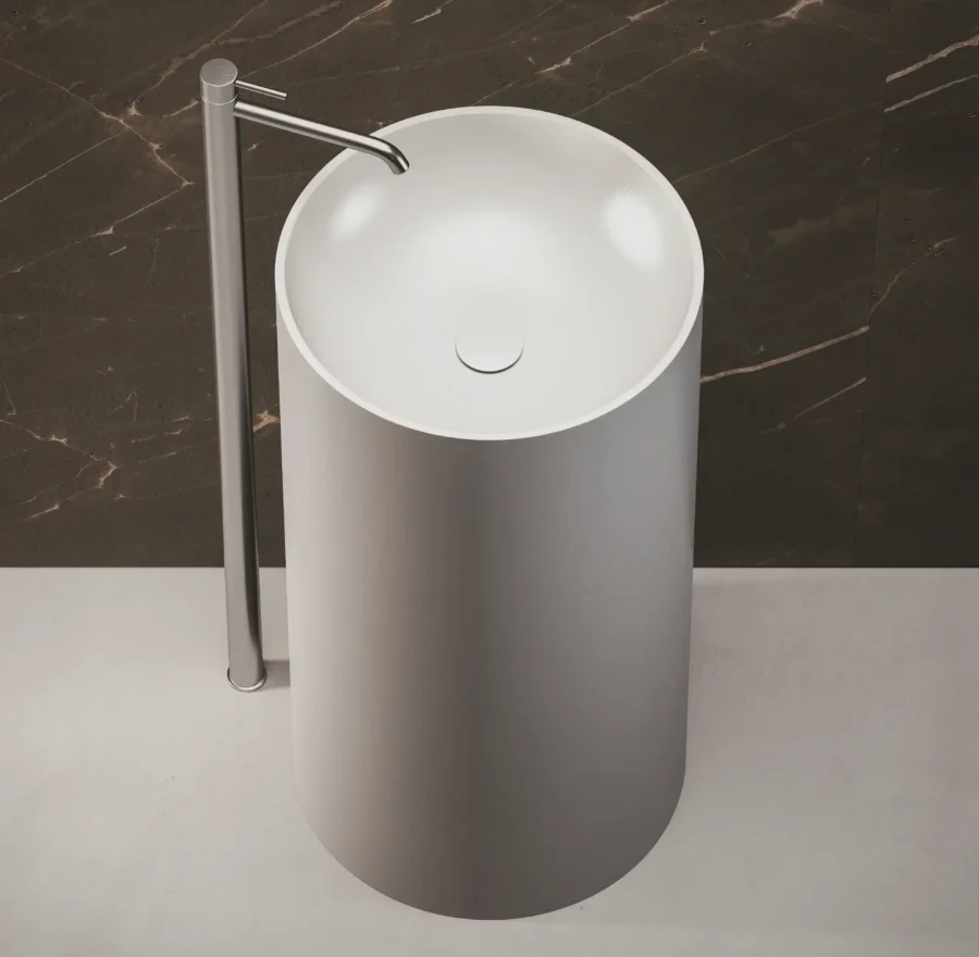 Lavabo freestanding cilindrico grigio chiaro con rubinetto cromato, visto dall’alto su fondo marmoreo scuro.