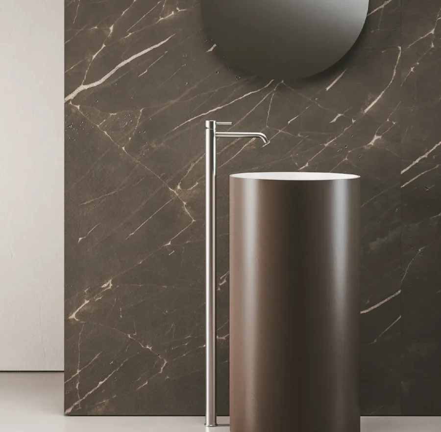 Lavabo freestanding cilindrico marrone con rubinetto a colonna cromato su sfondo effetto marmo scuro.