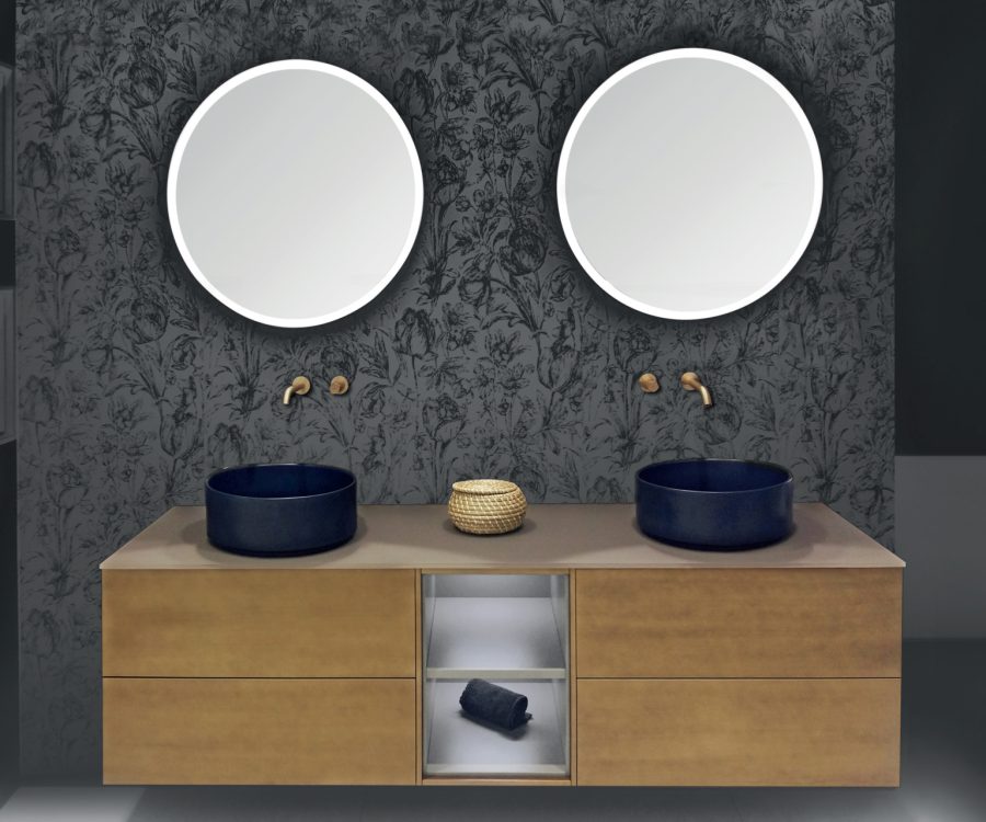 : Mobile bagno sospeso in finitura bronzo con doppio lavabo tondo nero opaco, due specchi rotondi retroilluminati e sfondo con carta da parati floreale scura.