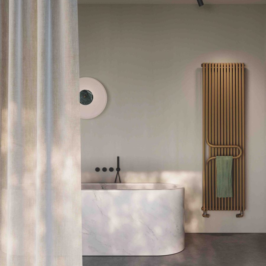 Bagno con vasca freestanding effetto marmo e radiatore verticale dorato con porta asciugamano integrato.
